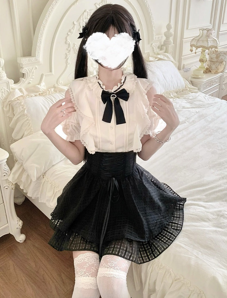 A person wearing a Lolita-style outfit consisting of a cream ruffled blouse with a black bow, a black pleated skirt, and white lace socks, posing on a white bed.
