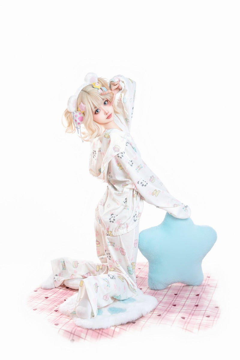 A young girl with blonde pigtails poses in a cozy white pajama set with pastel cartoon prints, kneeling on a pink plaid blanket next to a light blue star-shaped pillow against a solid white background.