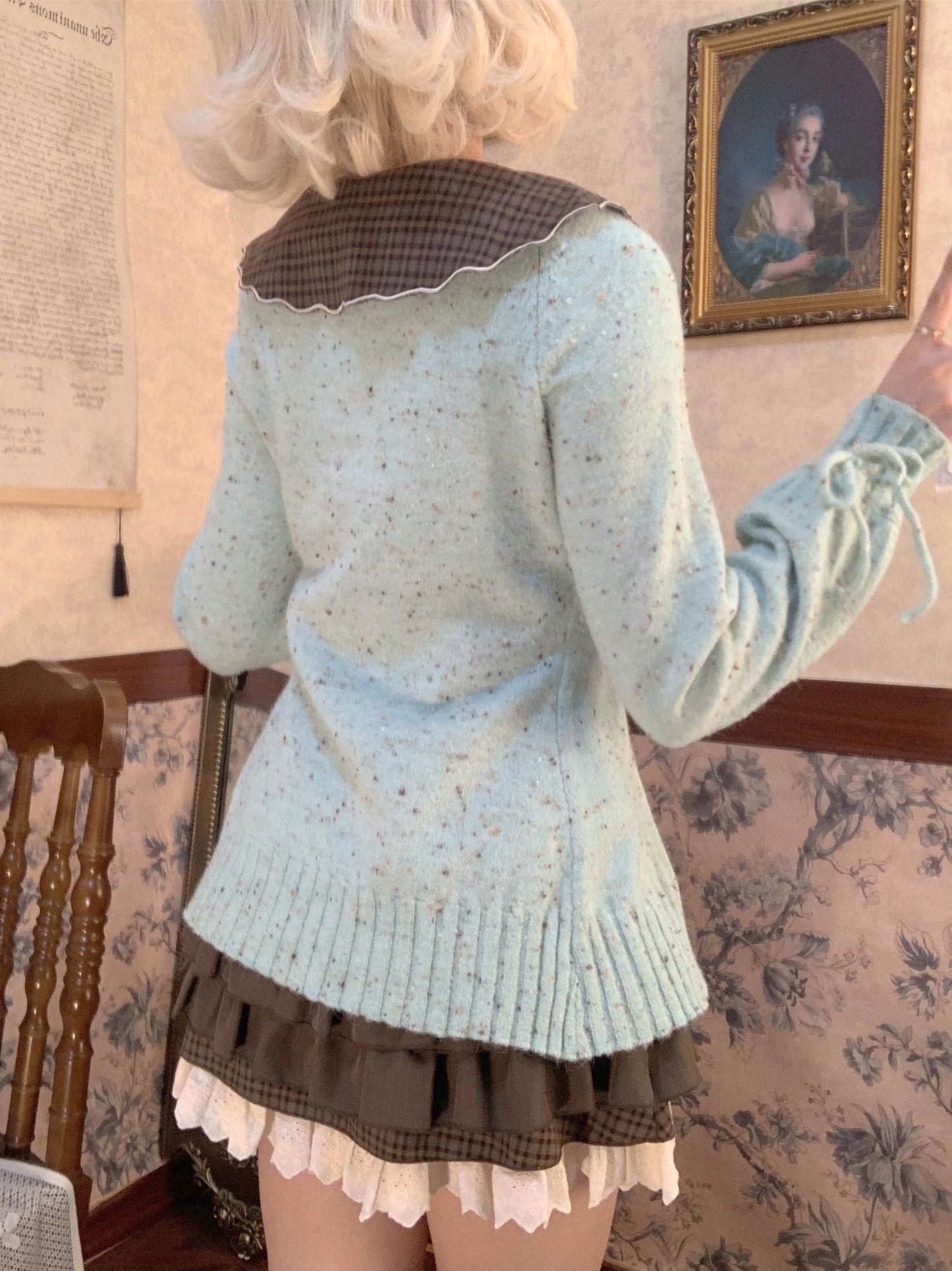 Person wearing a light blue knitted sweater and brown skirt in a room with floral wallpaper.