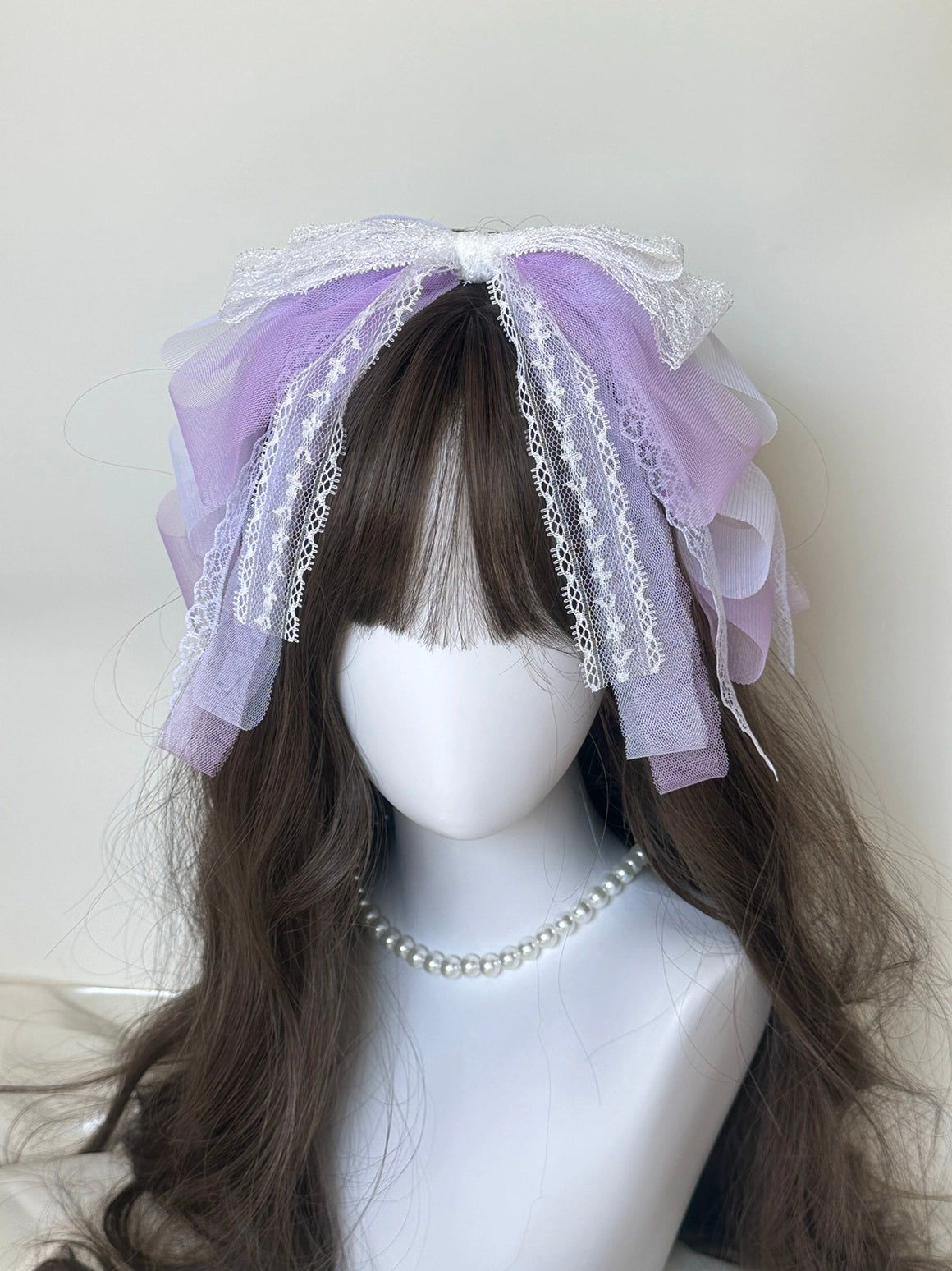 A lavender and white lace hair bow headpiece displayed on a white mannequin head with long brown hair and a pearl necklace.