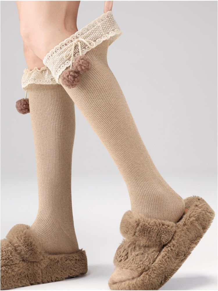 Beige knee-high socks with lace and pom-poms paired with fuzzy slippers on a light gray background