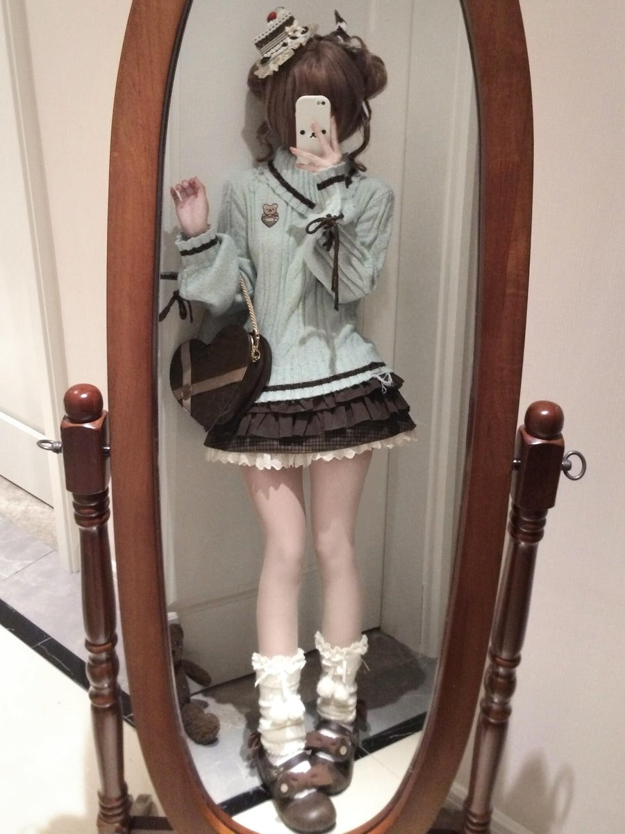 A person taking a mirror selfie wearing a light sage green ribbed sweater, a dark brown pleated mini skirt with white lace trim, white ruffled socks, and brown shoes. The setting is indoors in front of a large wooden-framed floor mirror.