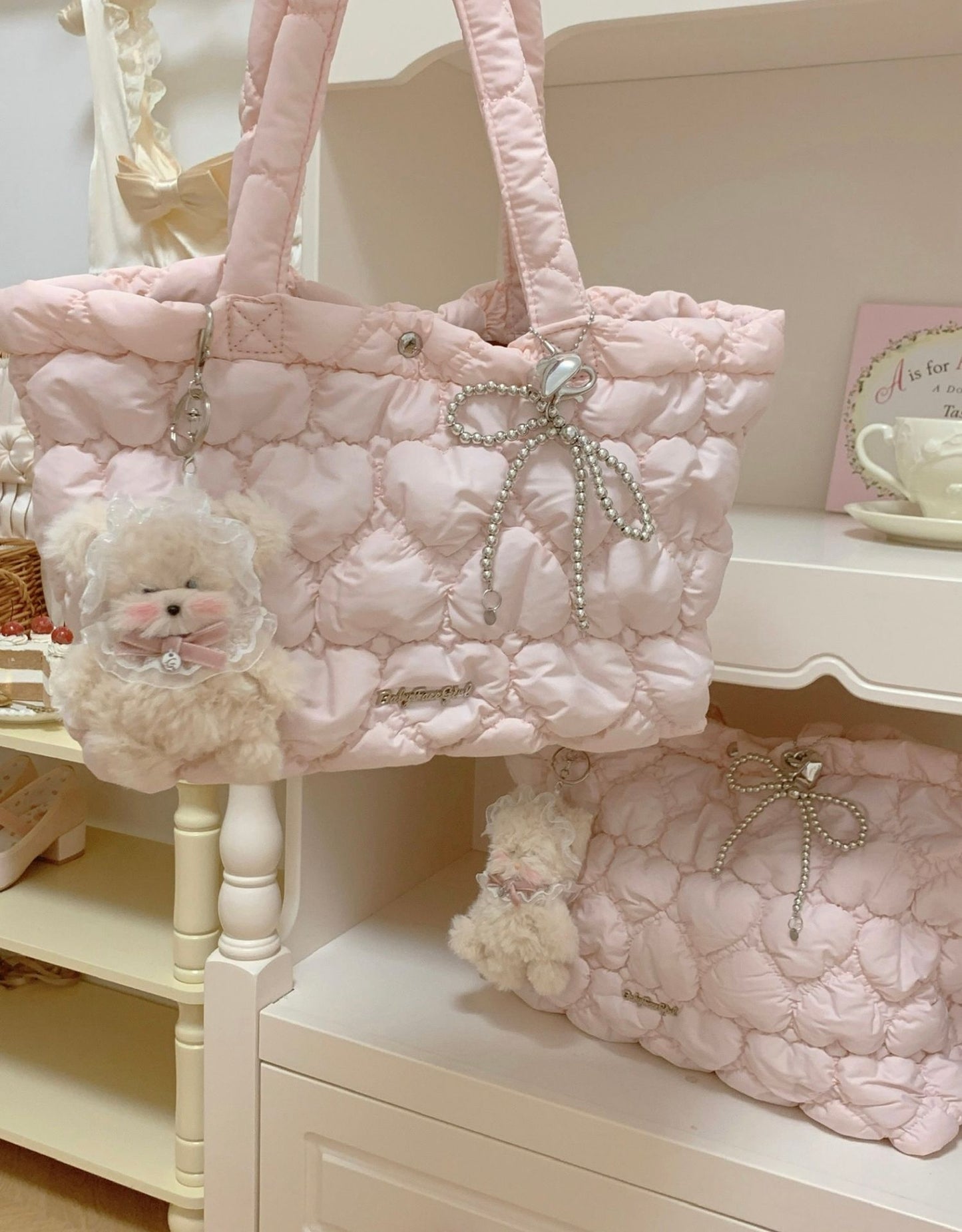 A light pink quilted puffer tote bag with pearl-beaded bow details and a fluffy cream plush charm, displayed on a white shelf alongside a matching pouch.