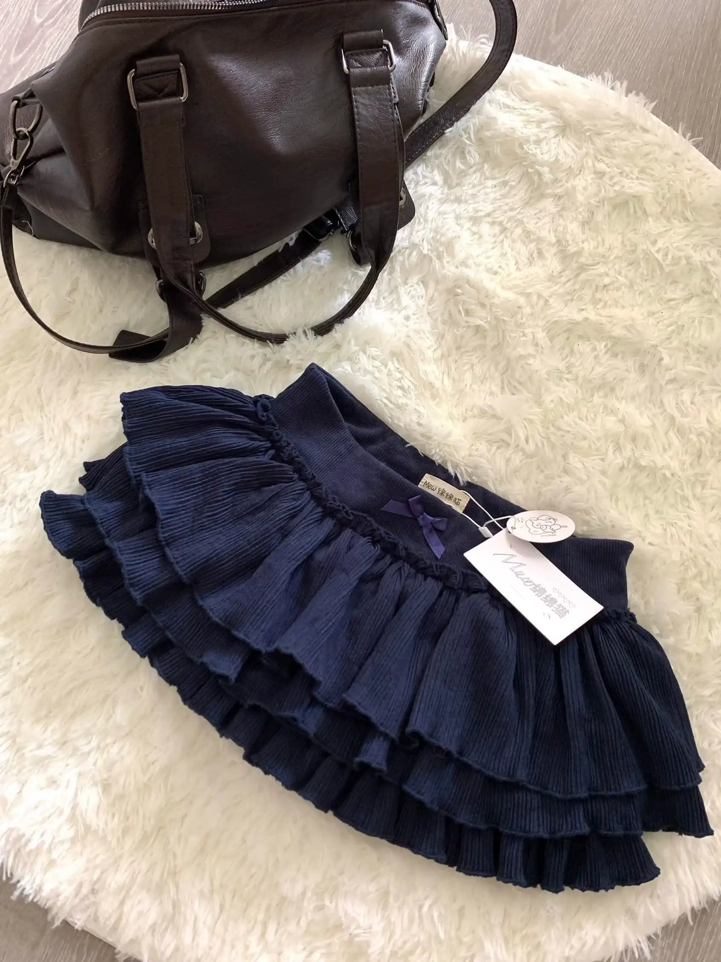 Navy blue ruffled skirt with a tag on a white surface next to a dark brown bag.