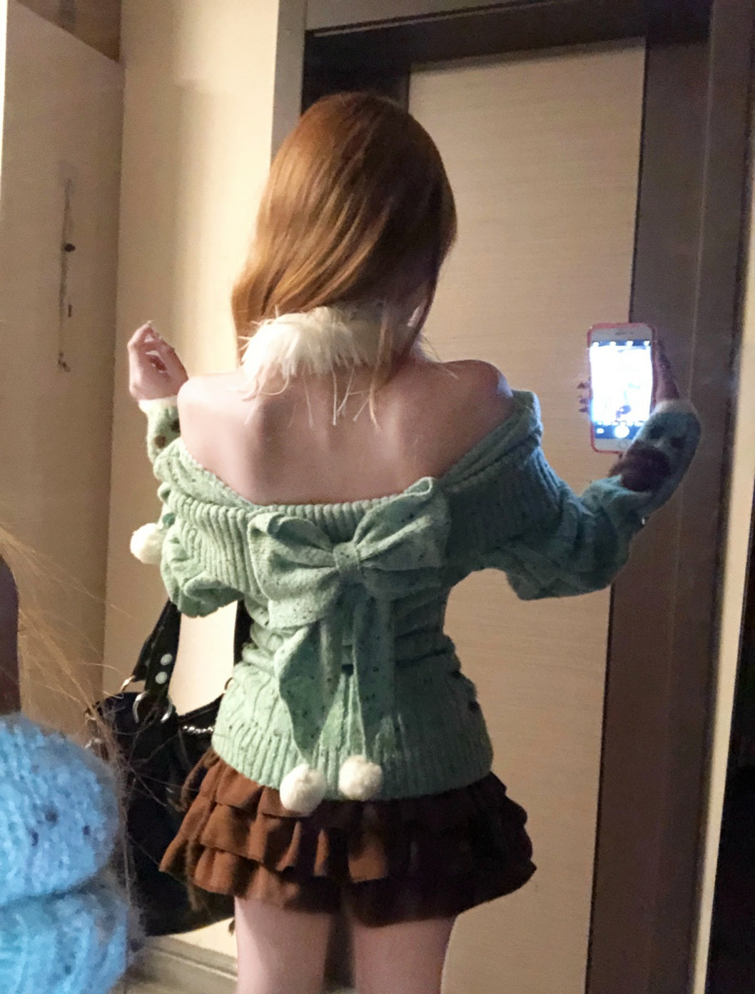 Person wearing a green sweater with a bow and brown skirt, taking a mirror selfie.
