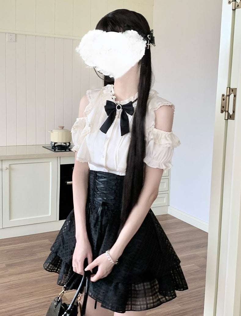 A person wearing a white cold-shoulder blouse with a large black bow at the collar and a black pleated skirt, standing indoors and holding a handbag. The face is obscured by a white heart-shaped blur.