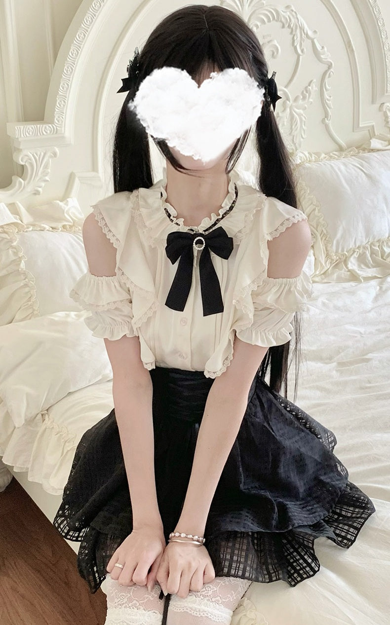 A person sitting on a bed wearing a cream and black Lolita-style outfit with a ruffled blouse, black bow, and black skirt, against a white bedroom background.