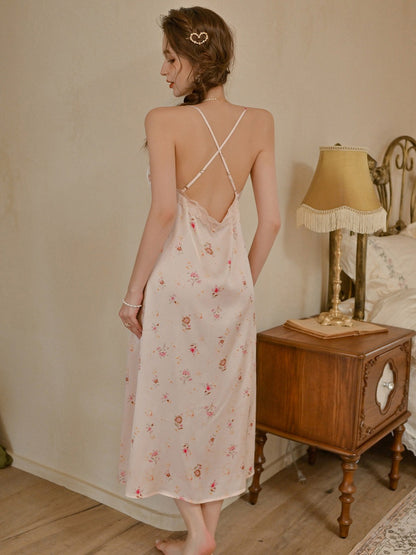 A woman stands barefoot in a bedroom wearing a light pink satin slip dress with a small floral print and a crisscross open back. A wooden nightstand with a lamp is visible beside her.