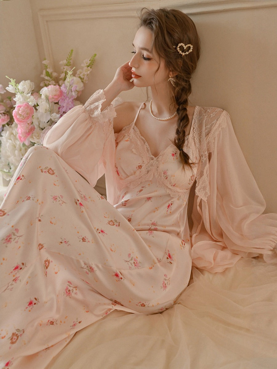 A woman wearing a light pink floral nightgown and matching sheer long sleeves, posed in a softly lit room with flowers and a cream-colored wall in the background.