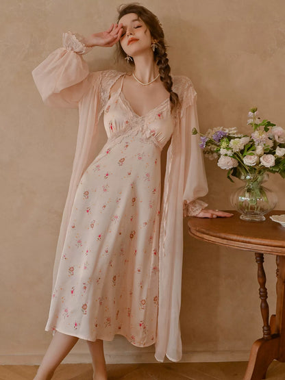 A woman models a light pink floral slip dress with a matching sheer long-sleeve cardigan, standing in a warm, neutral-toned room next to a wooden table with a vase of flowers.