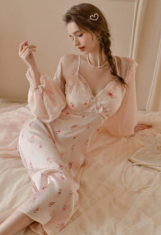 A woman wearing a light pink floral satin slip dress with a matching sheer long-sleeve overlay, sitting on a bed with a pearl necklace and a pearl handbag nearby.