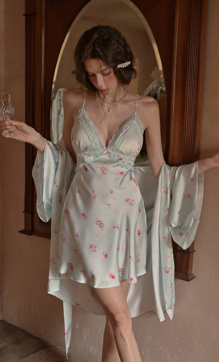 A woman models a light sage green satin nightgown with a pink floral print, paired with a matching long-sleeve robe, set against a warm-toned wooden archway background.