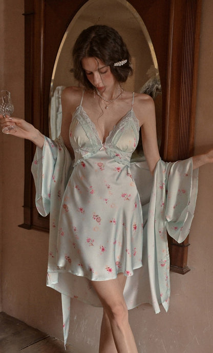 A woman models a light sage green satin nightgown with a pink floral print, paired with a matching long-sleeve robe, set against a warm-toned wooden archway background.