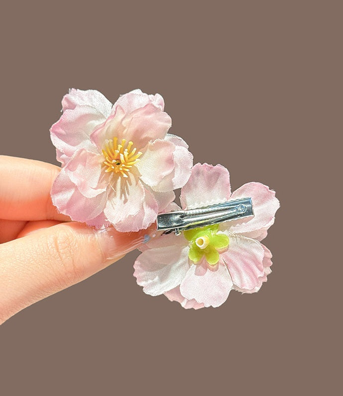 A close-up of a hand holding a silver hair clip decorated with two light pink cherry blossom flowers and a small green center, against a solid taupe background.