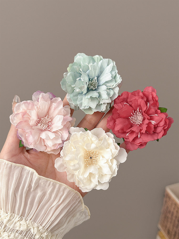 Four fabric peony-style flowers in pastel pink, mint green, cream white, and coral red, held in a hand against a plain taupe background.