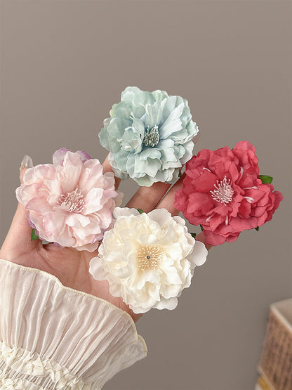 Four fabric peony-style flowers in pastel pink, mint green, cream white, and coral red, held in a hand against a plain taupe background.