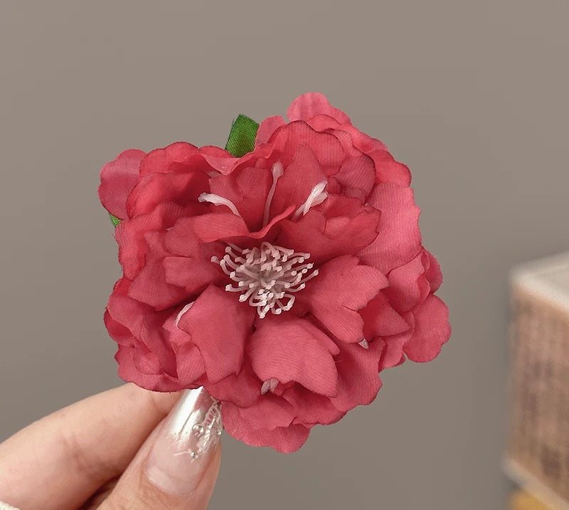 A close-up of a hand holding a large, deep pink peony-style flower with layered petals, a green stem, and a white center against a soft gray background.