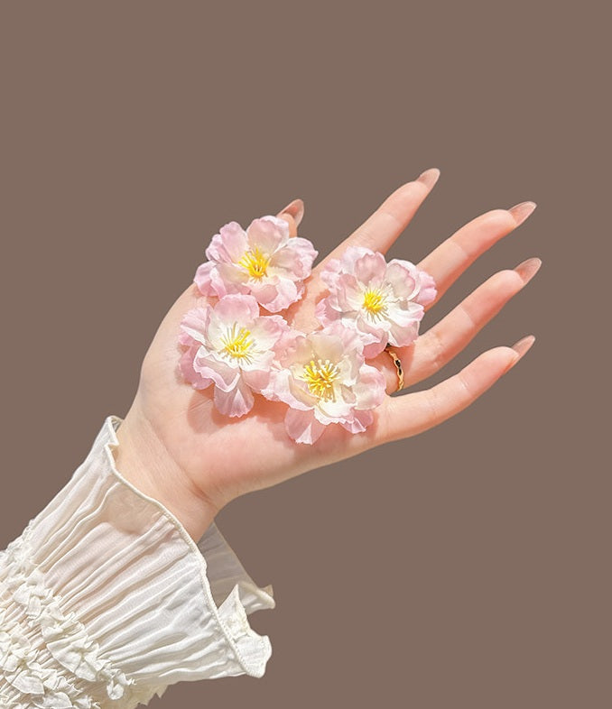 A hand wearing a white ruffled long-sleeve blouse holds four small artificial pink flowers with yellow centers against a solid taupe-brown background.