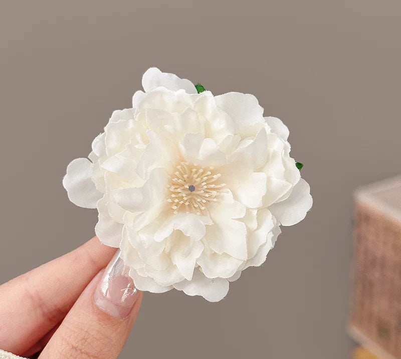 A hand holding a large white artificial flower accessory with layered petals and a cream-colored center, photographed against a soft neutral background.