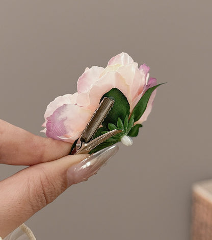 A close-up of a hand holding a small artificial flower hair clip. The clip features a pale pink and white peony-style flower with green leaves and a silver metal spring clip mechanism, shown against a neutral gray background.