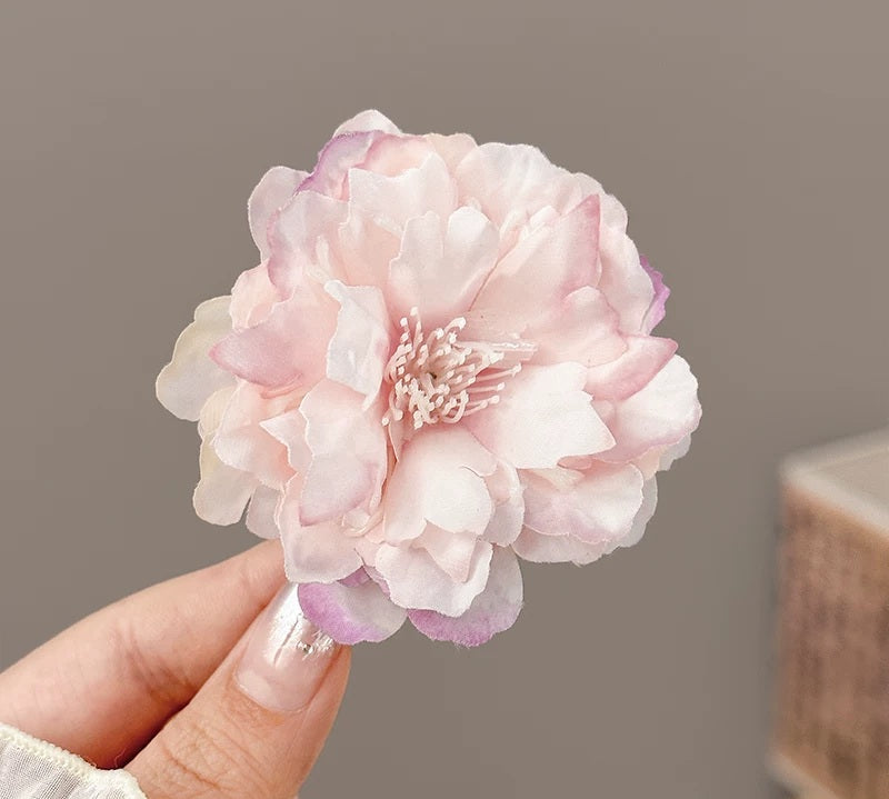 A hand holding a large artificial pink peony flower with layered petals and a pale center, shown against a soft gray background.
