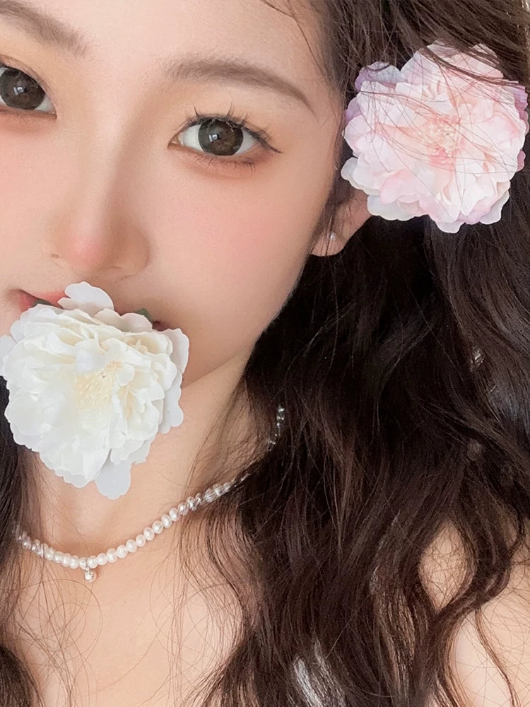 A close-up portrait of a woman with long dark brown hair, wearing a delicate pearl necklace and a large pale pink flower hair accessory on one side of her head, with a white flower held near her mouth.