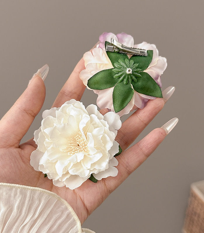 Two flower-shaped hair clips held in a hand: one white peony with a cream center and one layered pale pink and white flower with green leaves and a small rhinestone accent, shown against a soft taupe background.