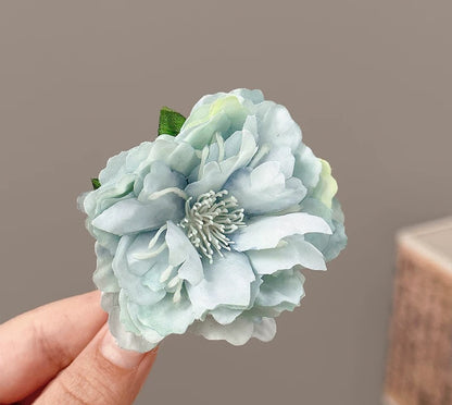 A pale blue and white artificial peony flower with green leaves, held by a person's fingers against a neutral gray background.