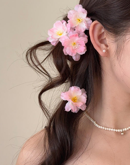A close-up of a woman's hair styled with three pink cherry blossom flower hair clips, a pearl stud earring, and a pearl necklace against a soft beige background.