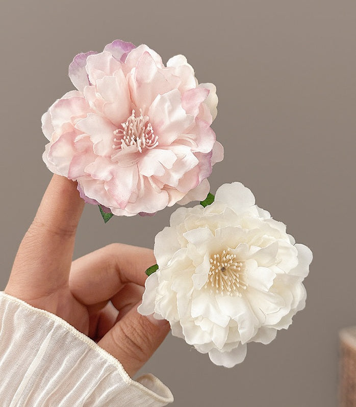 A hand wearing a cream-colored sleeve holds two large artificial peony-style flowers against a plain taupe background. One flower is pale blush pink with soft purple shading, and the other is creamy white with pale gold centers and small green stems.