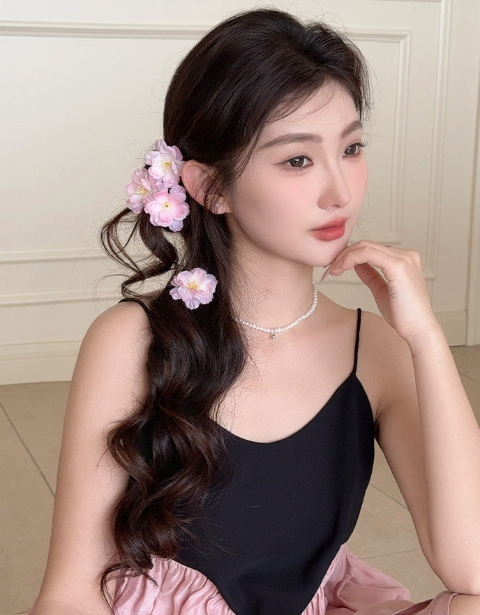 A woman with long, dark, wavy hair models pink fabric flower hair clips. She is wearing a black camisole top and a light pink skirt, posing indoors against a cream-colored paneled wall.