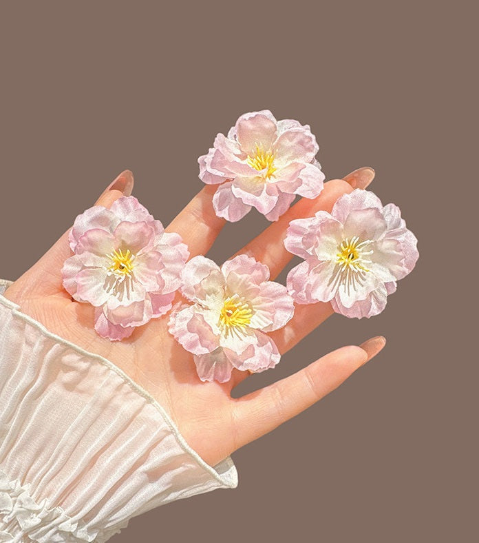 Four light pink cherry blossom flower clips held in a hand against a solid taupe background.