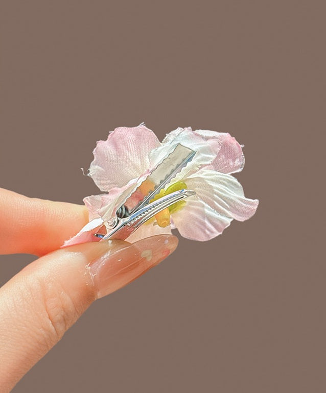 A close-up of a hand holding a small flower hair clip against a solid taupe-brown background. The clip features light pink and white fabric petals with a small yellow accent in the center and a silver metal spring.