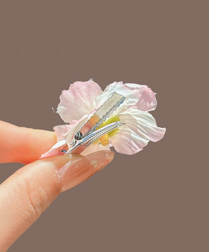 A close-up of a hand holding a small flower hair clip against a solid taupe-brown background. The clip features light pink and white fabric petals with a small yellow accent in the center and a silver metal spring.