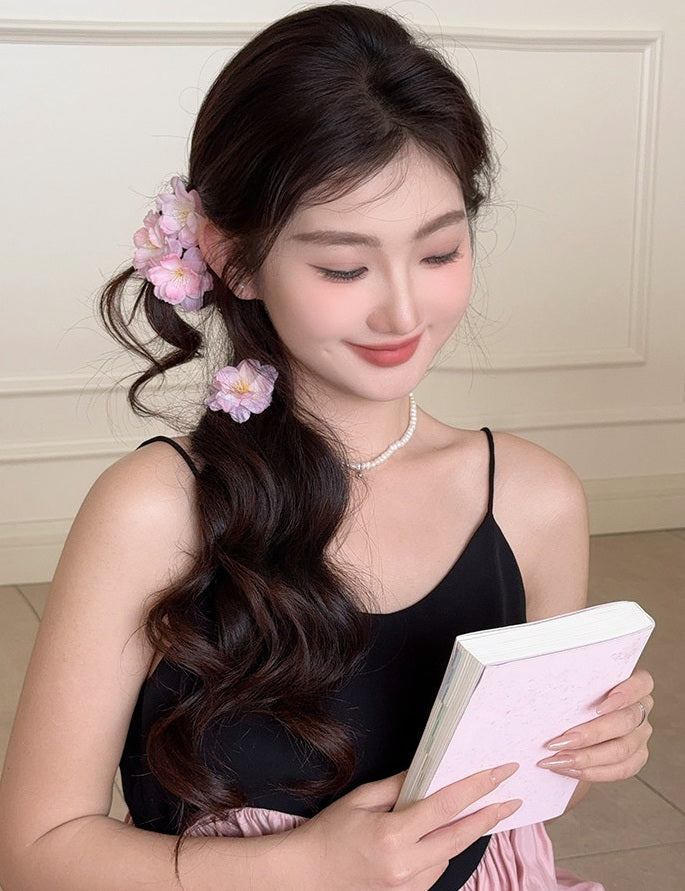 A woman with long, wavy dark brown hair wearing pink flower hair clips, a black camisole, and a pearl necklace, holding a light pink notebook.