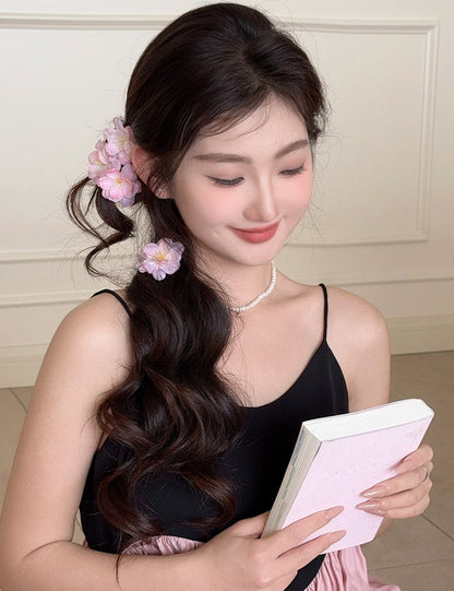 A woman with long, wavy dark brown hair wearing pink flower hair clips, a black camisole, and a pearl necklace, holding a light pink notebook.
