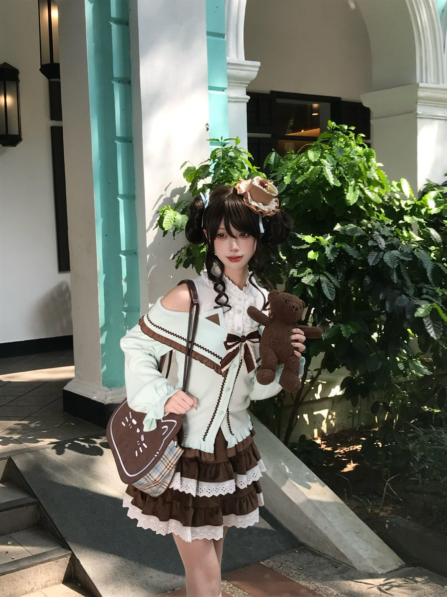 A model wearing a detailed Lolita-style outfit consisting of a cream and brown cardigan, a brown and cream pleated skirt, and a matching bag, holding a brown teddy bear in front of a building with green plants.