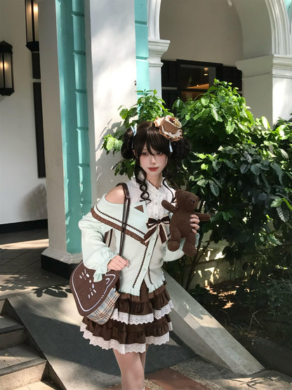 A model wearing a detailed Lolita-style outfit consisting of a cream and brown cardigan, a brown and cream pleated skirt, and a matching bag, holding a brown teddy bear in front of a building with green plants.