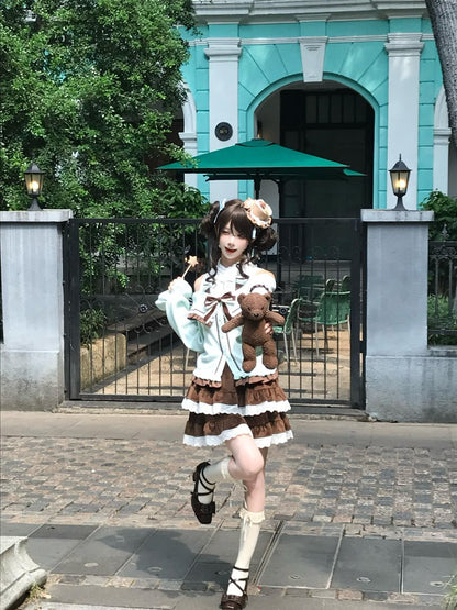 A woman posing outdoors in front of a teal building and black gate, wearing a brown and white tiered Lolita-style dress with a light blue cardigan, holding a brown teddy bear.