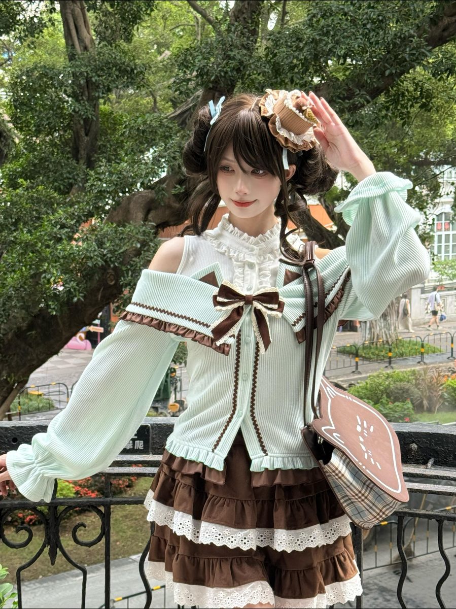 A model wearing a light mint green and brown Lolita-style outfit with lace details, a large bow, and a pleated skirt, standing outdoors in front of a tree.