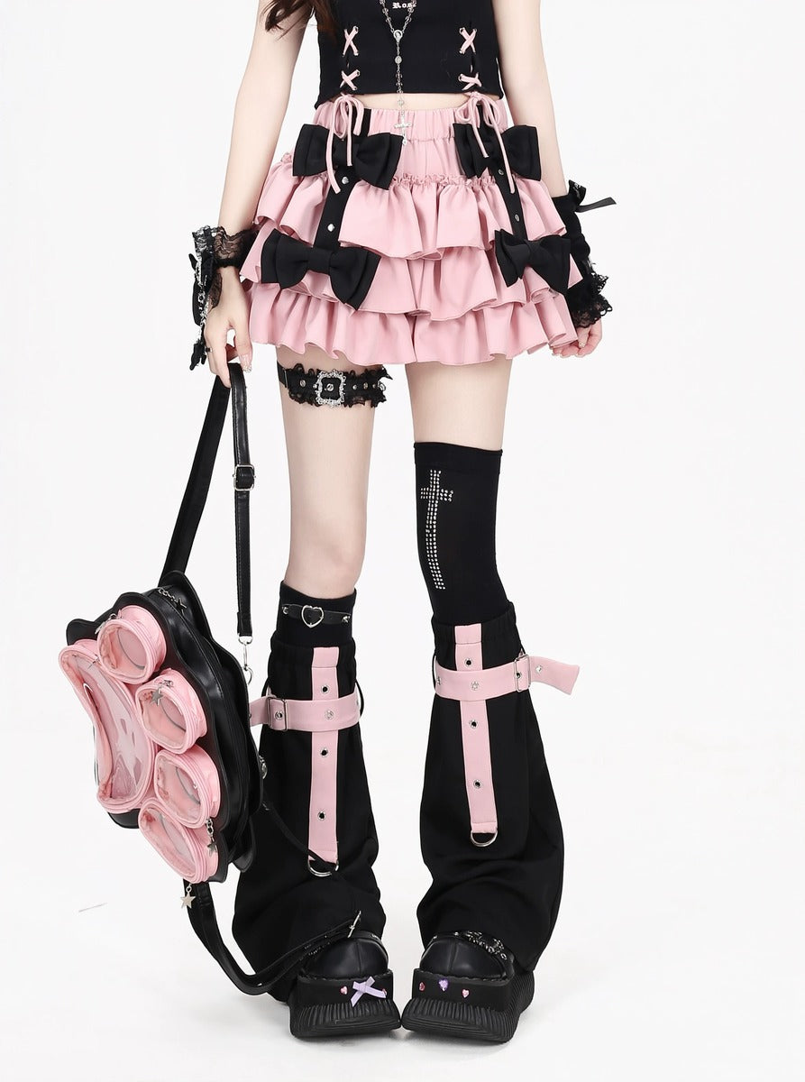 A model wearing a black and pink goth-style outfit, featuring a ruffled pink mini skirt with black bows, black thigh-high socks with pink straps, and a black backpack with pink flower details, set against a plain white background.