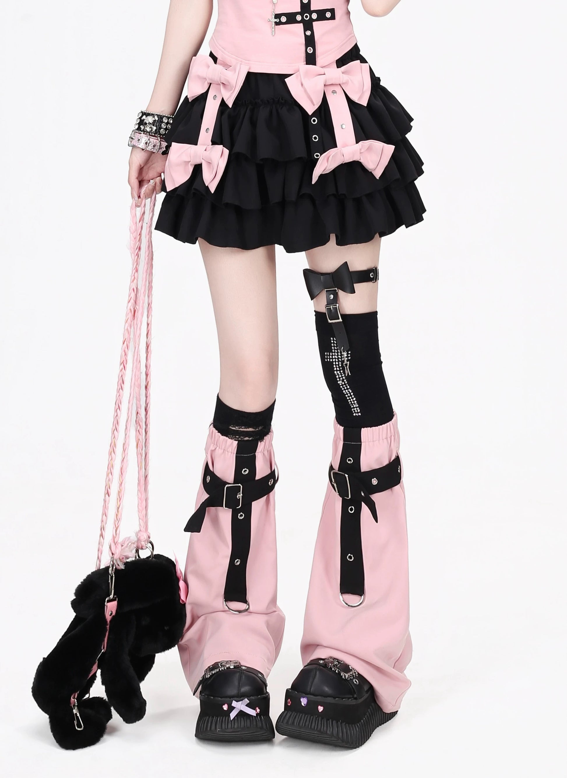 A person wearing a pink and black goth-style outfit with a tiered ruffled skirt, large pink bows, matching pink leg warmers with black straps, black knee socks, and black platform shoes. A small black fuzzy handbag with a pink strap is also shown against a plain white background.