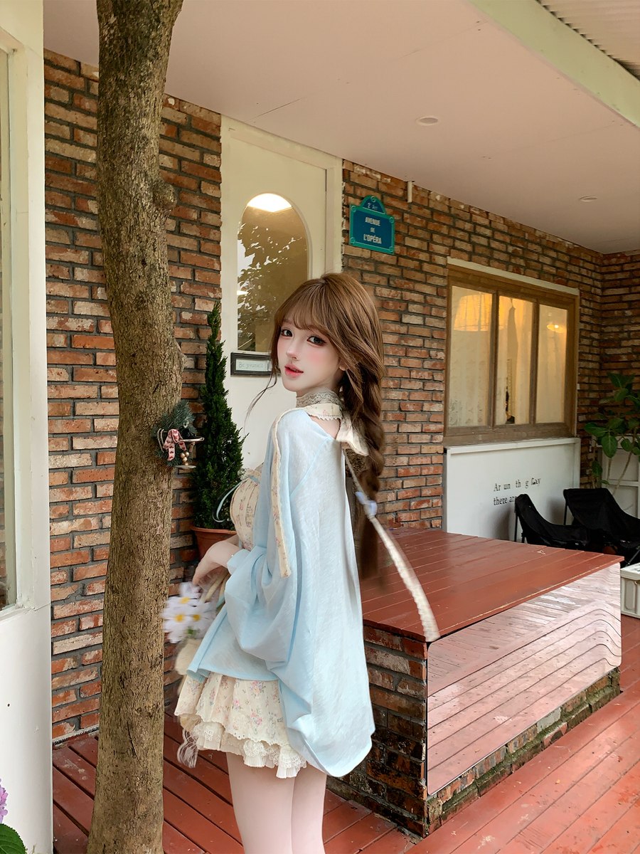 Woman in a light blue dress standing in front of a brick wall with a tree and wooden bench.