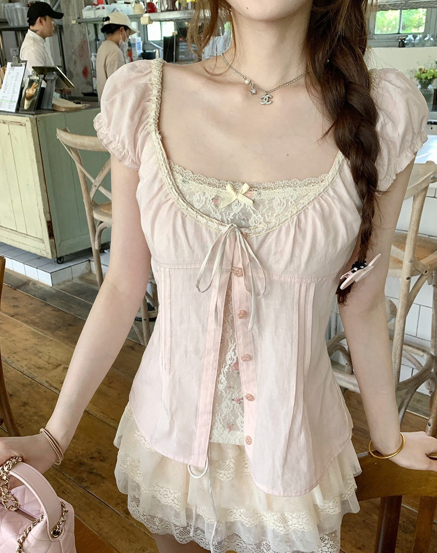 A woman wearing a pale pink short-sleeve lace-trim top with a square neckline, ruffled trim, and front buttons, photographed in a cafe setting.