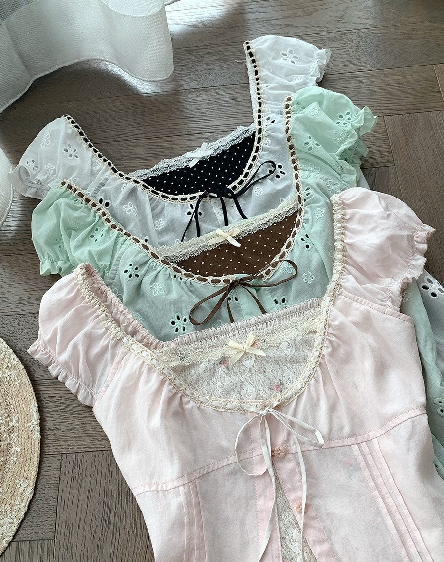 Three pastel lace-trimmed ruffled top pieces laid flat on a wood floor, shown in black-and-white polka dot, mint green, and pale pink, each with a bow detail.