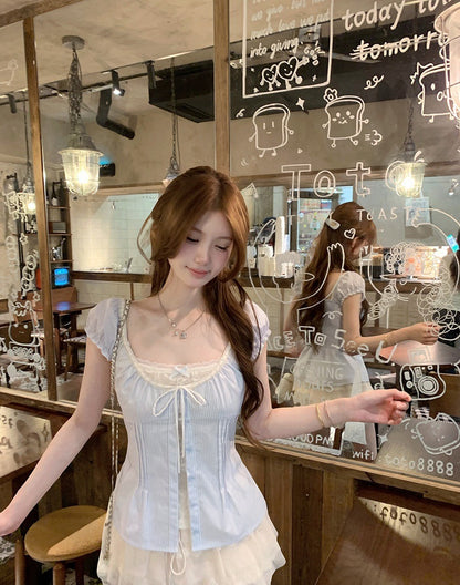 A woman wearing a light blue lace-trimmed corset-style top with short sleeves and a ruffled hem, standing in a cafe in front of a glass window with cartoon drawings on it.
