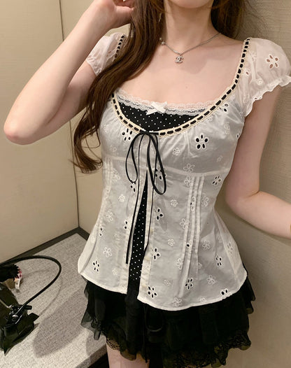 A woman wearing a white eyelet lace corset-style top with black polka-dot trim and a black tie detail, paired with a black ruffled skirt, photographed indoors against a beige wall.