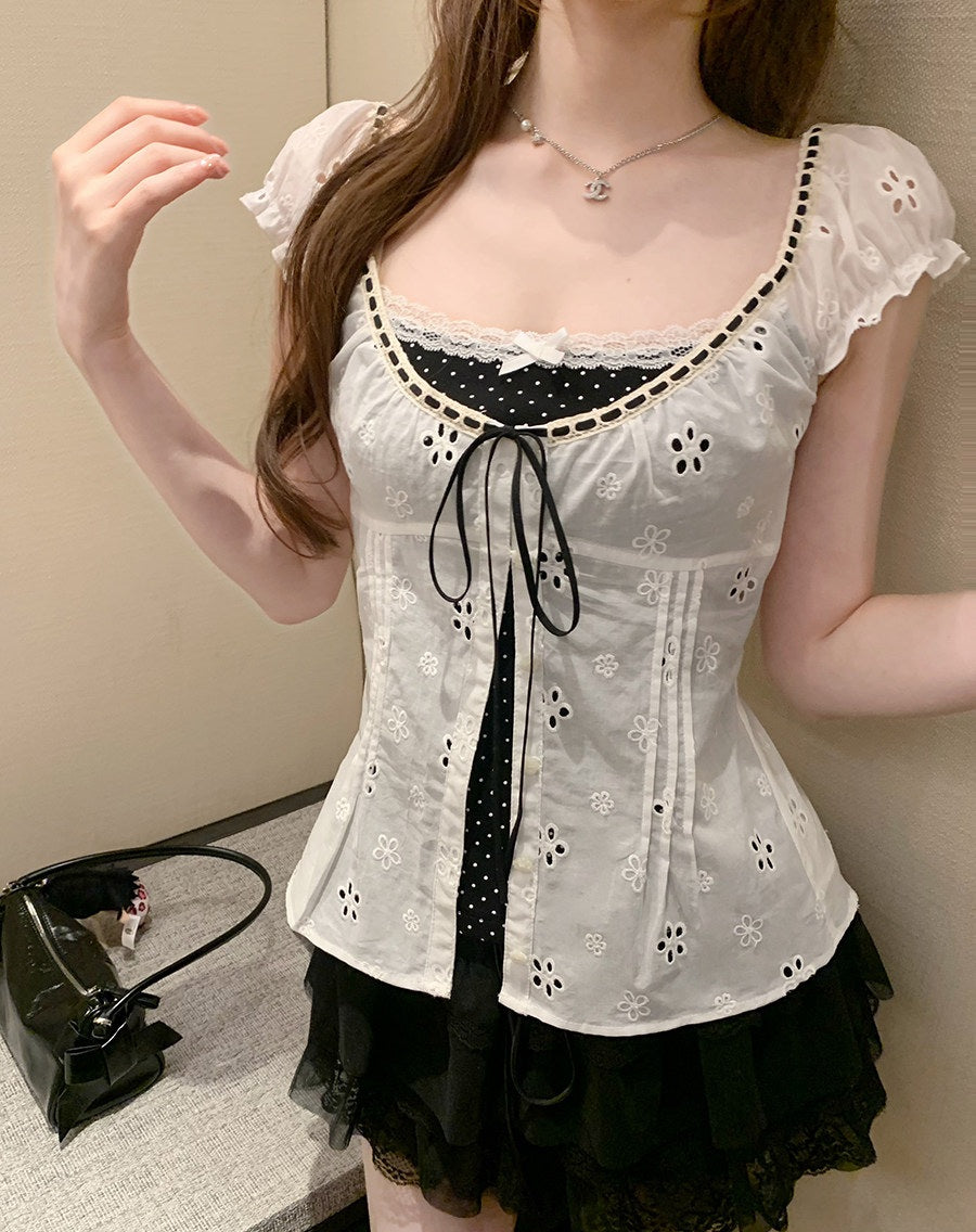 A woman wearing a white eyelet lace corset-style top with black polka-dot trim and a black ruffled skirt, standing indoors against a beige wall.