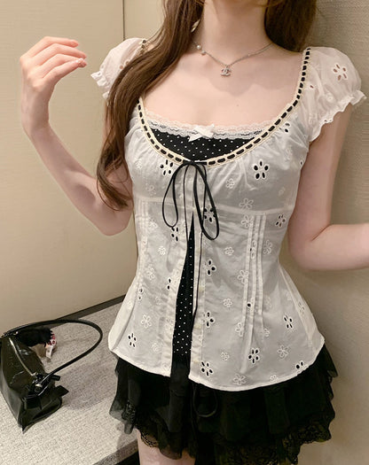 A woman wearing a white eyelet lace corset-style top with black polka-dot trim and a black ruffled skirt, standing indoors against a beige wall.