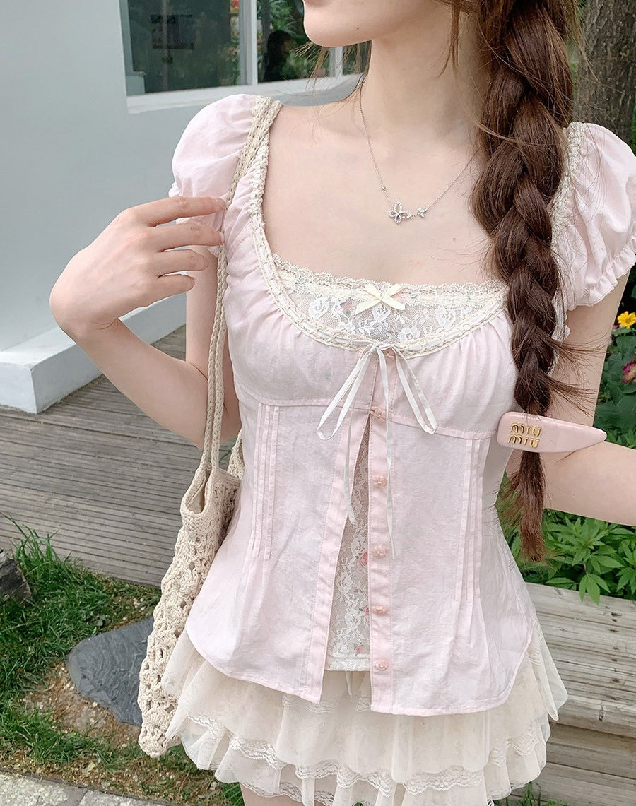 A woman wearing a pale pink short-sleeve lace-trim blouse with a bow tie detail and a matching ruffled skirt, photographed outdoors near a wooden fence and greenery.